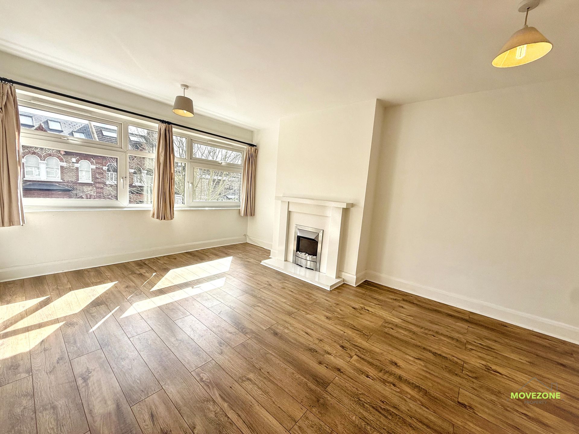 Empty living room with wood floors, large windows, and a white fireplace in a bright corner.
