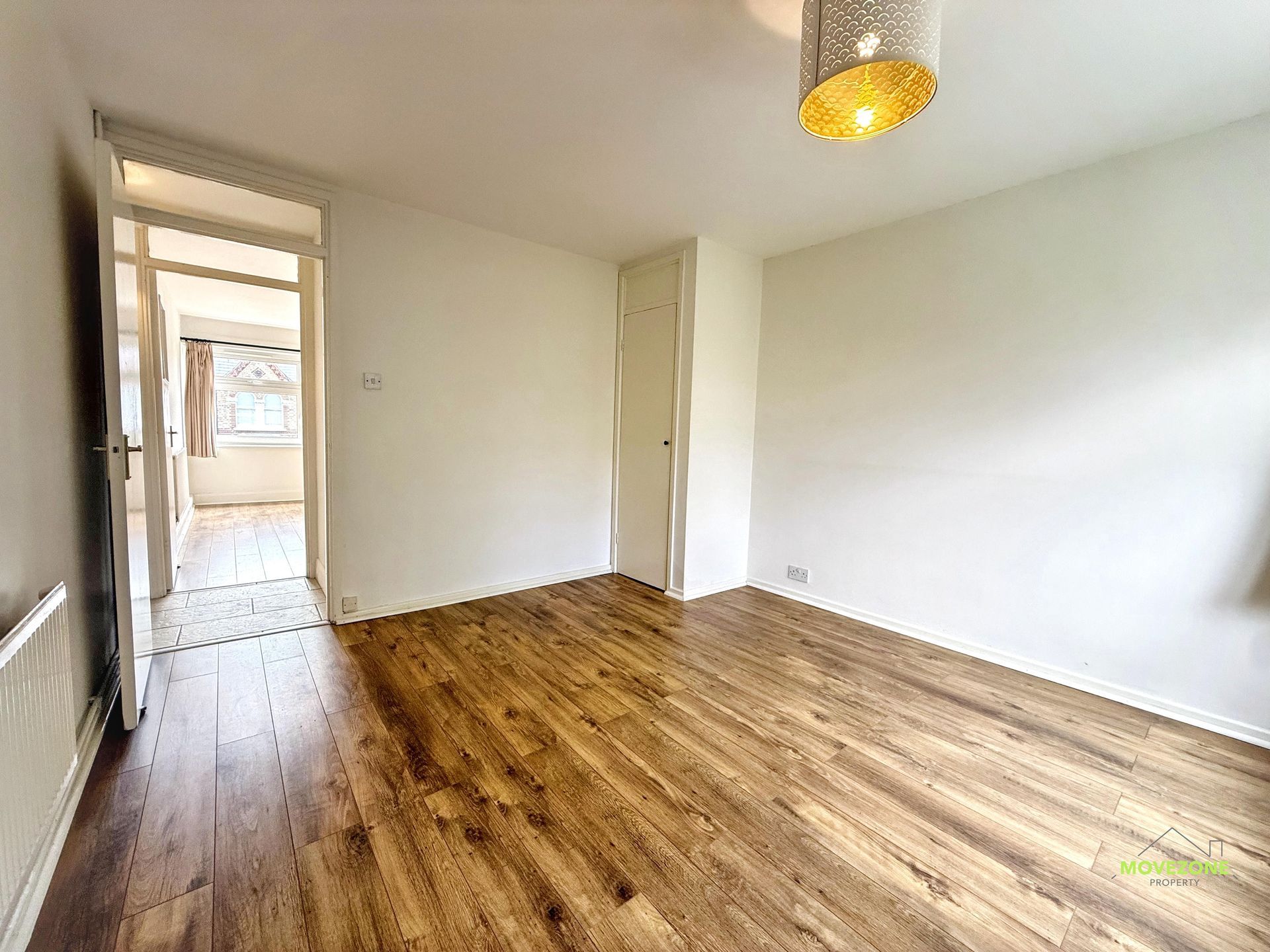 Empty room with wood floors, white walls, and a doorway to a hall, lit by a hanging ceiling lamp.
