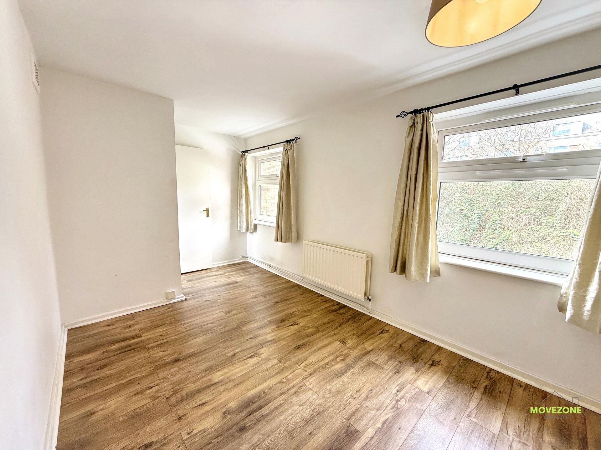Empty unfurnished room with wood floors, white walls, and two windows with curtains.