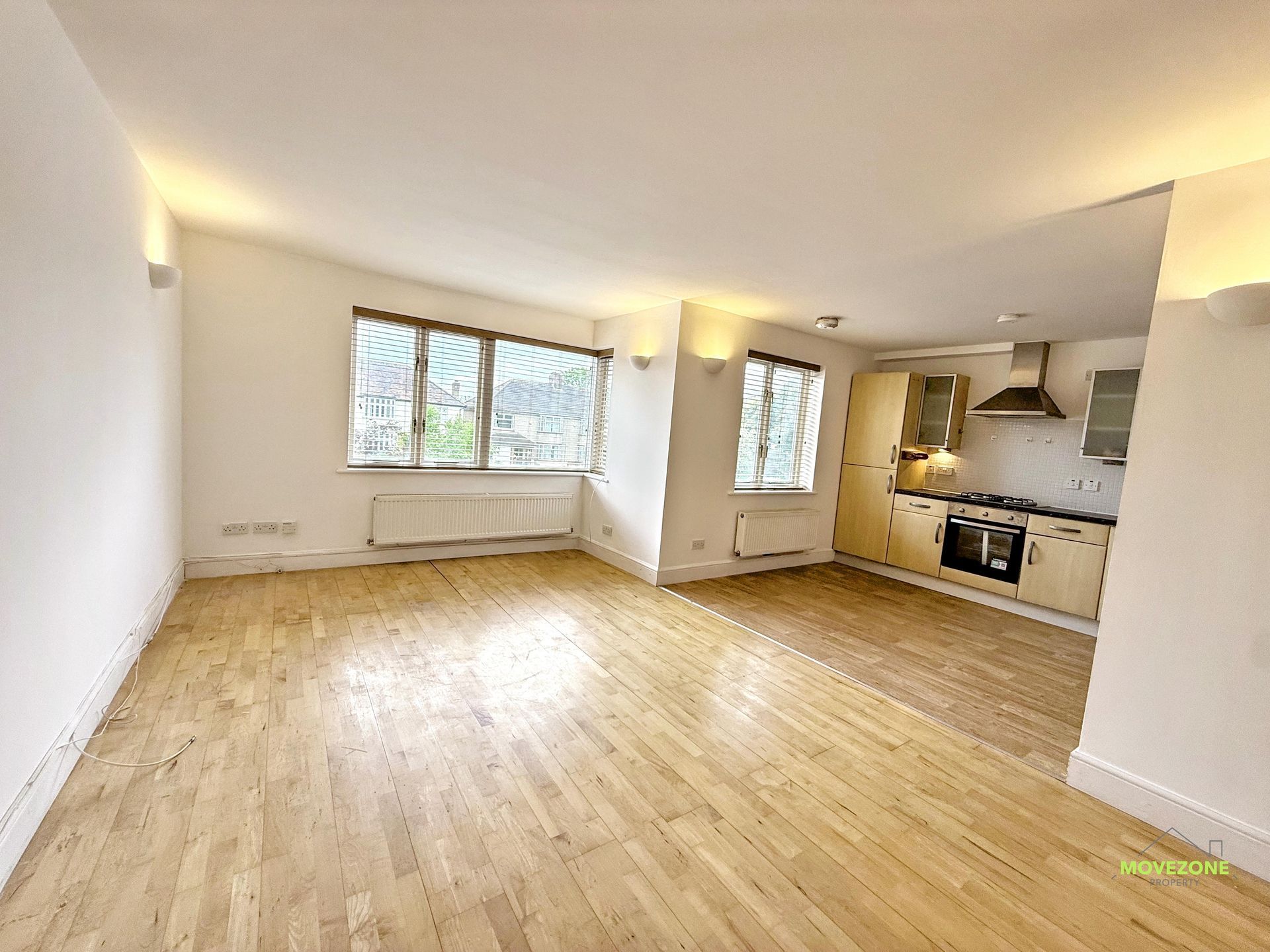 Empty open-plan living room with wood floors, window, radiator, and small kitchen area.