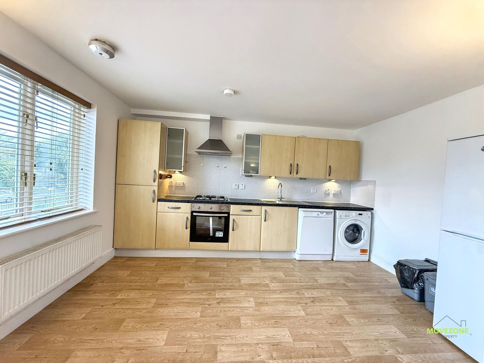 Empty modern kitchen with light wood cabinets, stainless range hood, window blinds, and a washing machine.