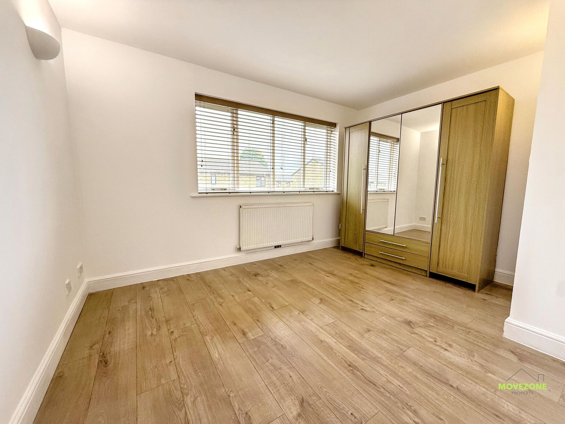 Empty bedroom with wood flooring, window blinds, radiator, and mirrored wardrobe.