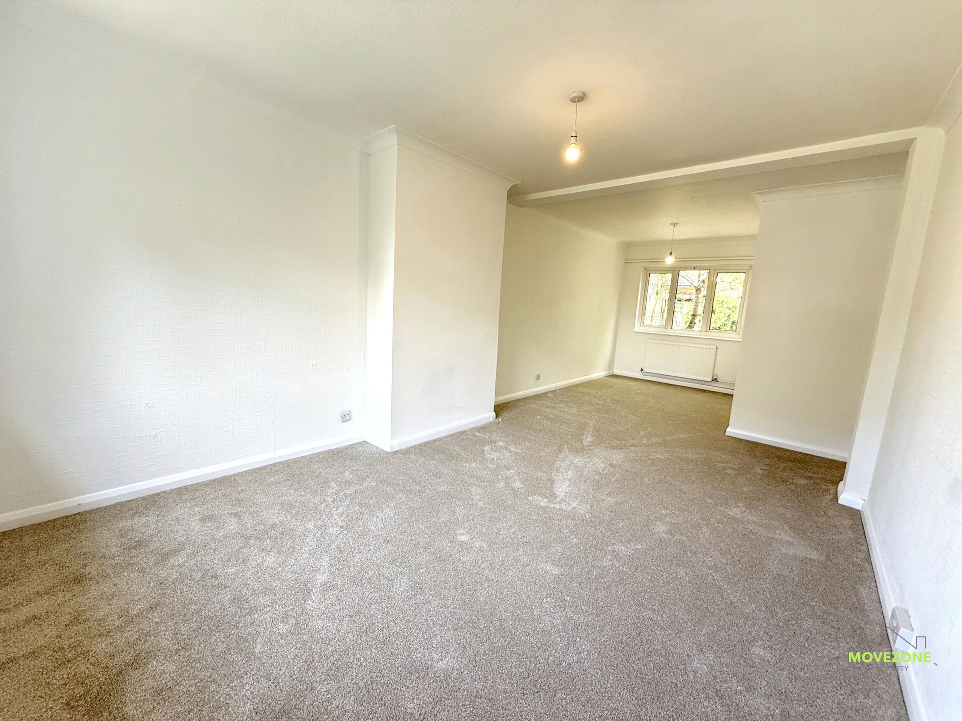 Empty beige-carpeted living room with white walls and a bright window opening to another room