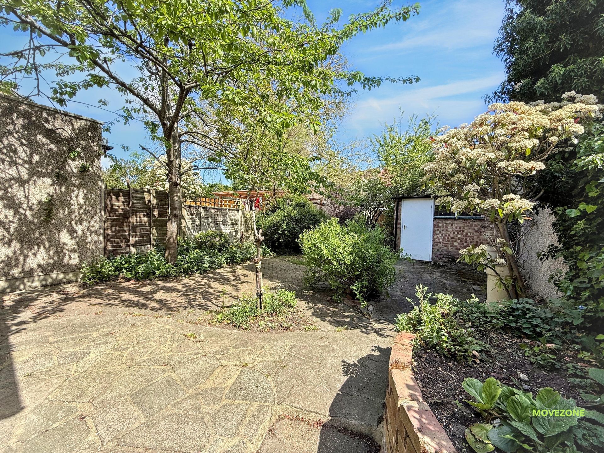 Stone patio garden with trees, shrubs, and a white gate under a blue sky