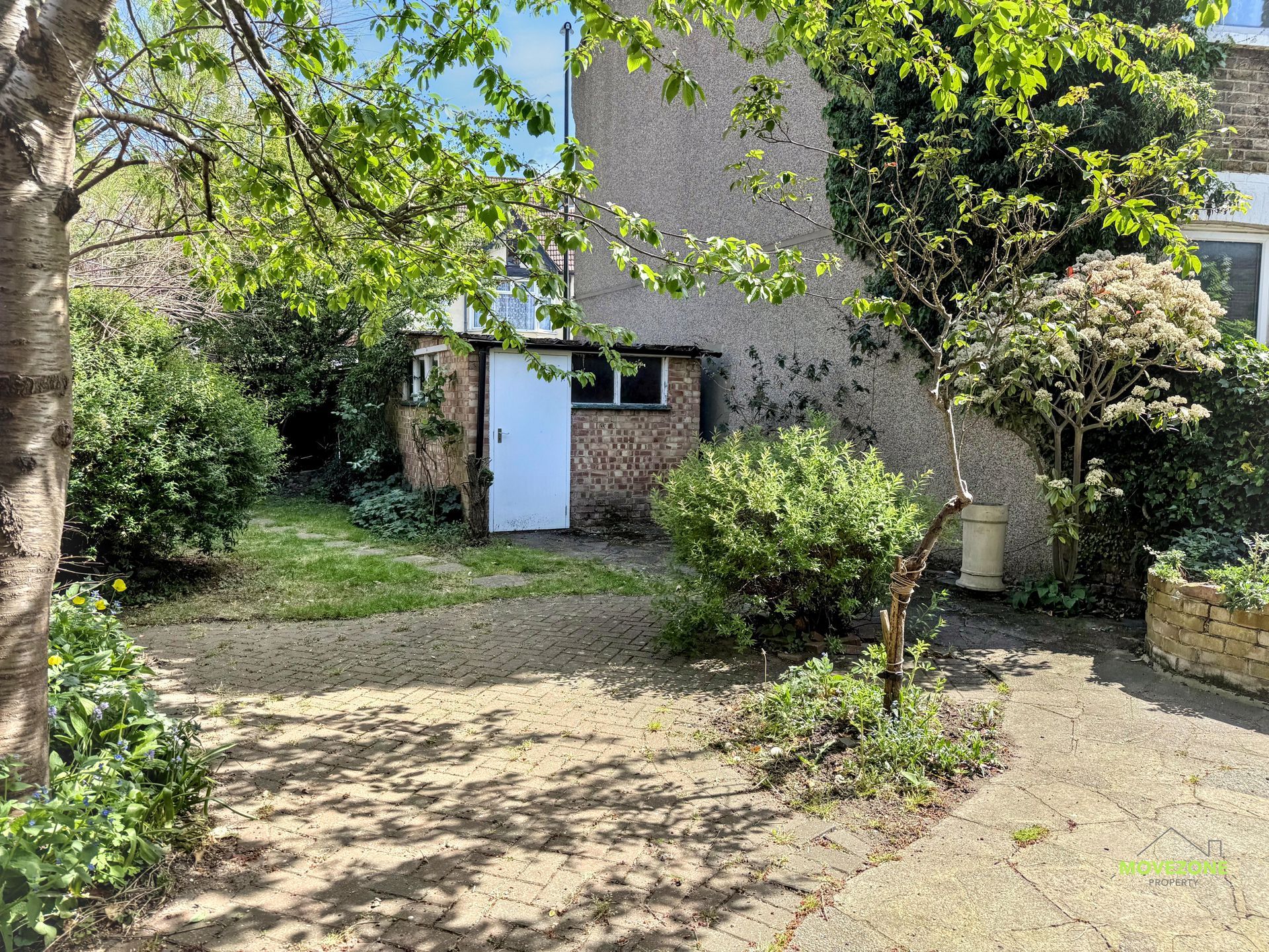 Small courtyard with trees, shrubs, and a white door on a stone building, lit by sunlight.