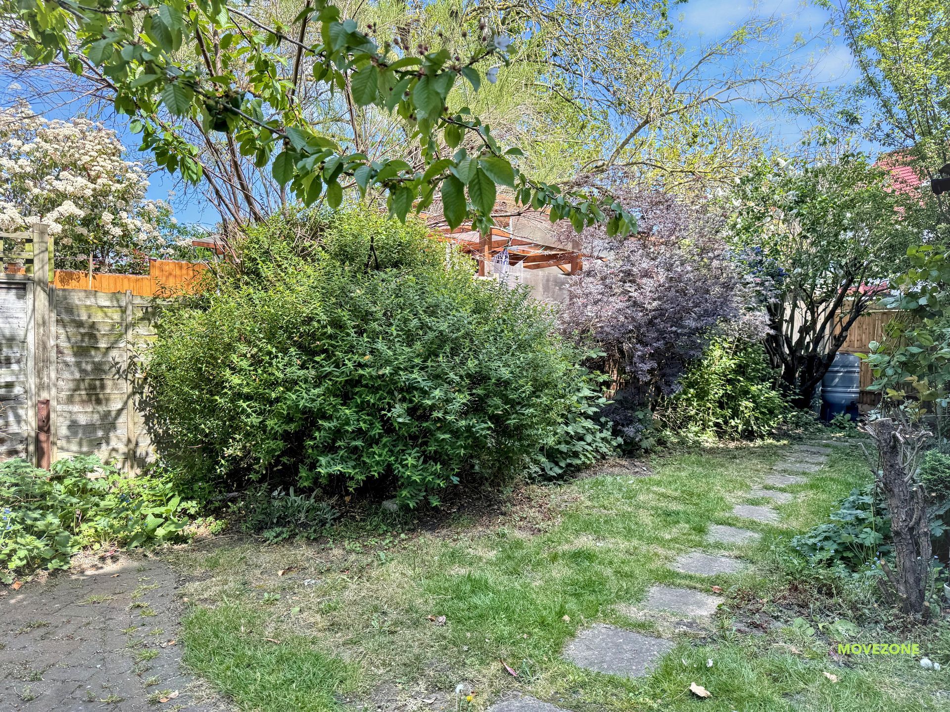 Garden path with stepping stones, lush bushes, and flowering trees under a blue sky.