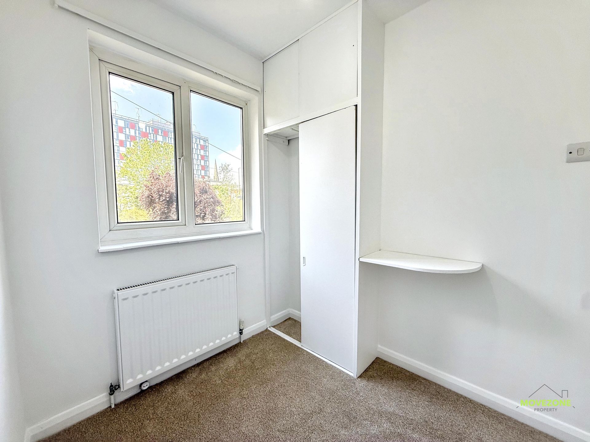 Small empty white room with a window, radiator, built-in wardrobe, and beige carpet.
