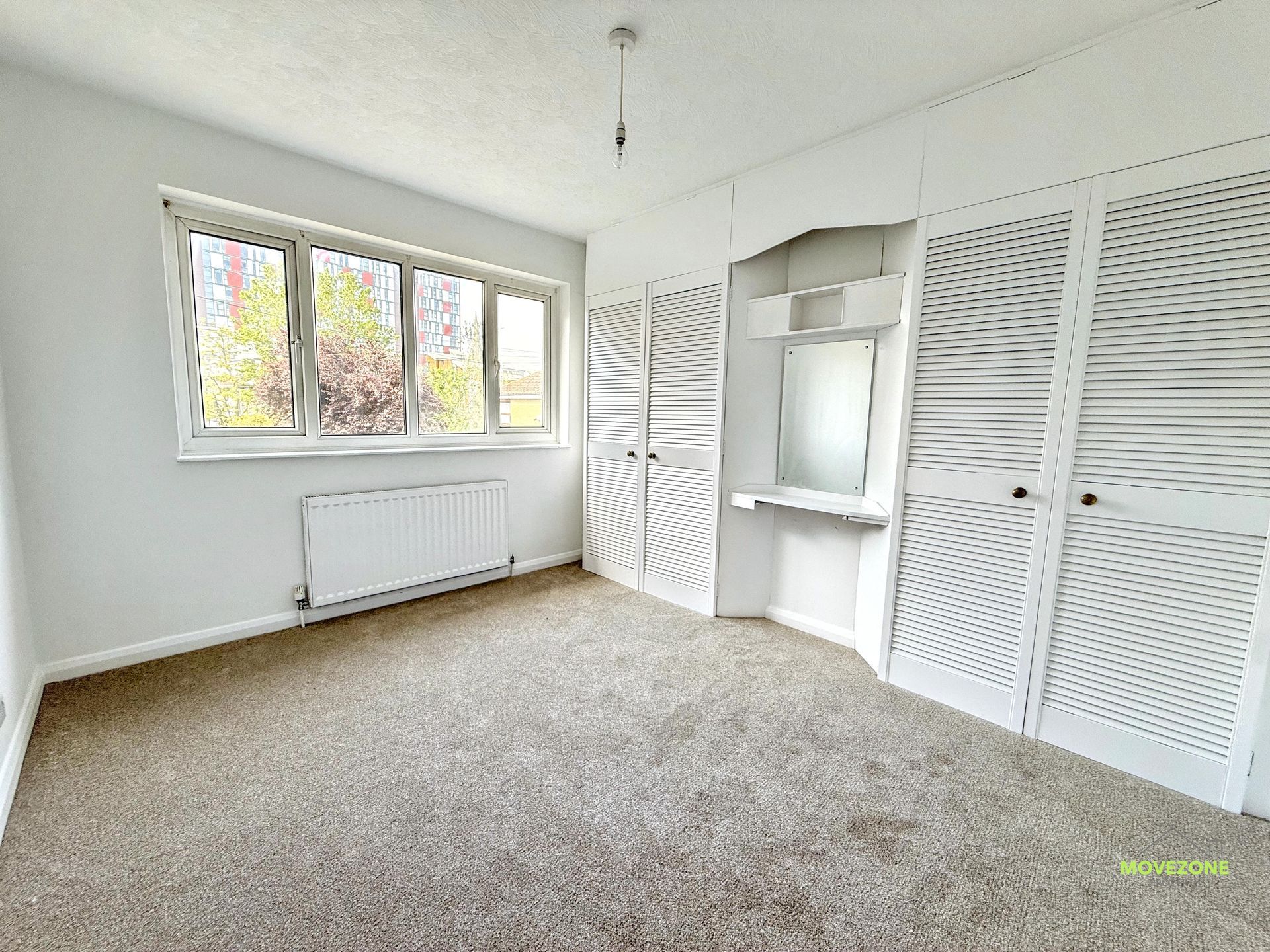 Bright empty bedroom with white walls, carpet, window, radiator, and built-in closets.