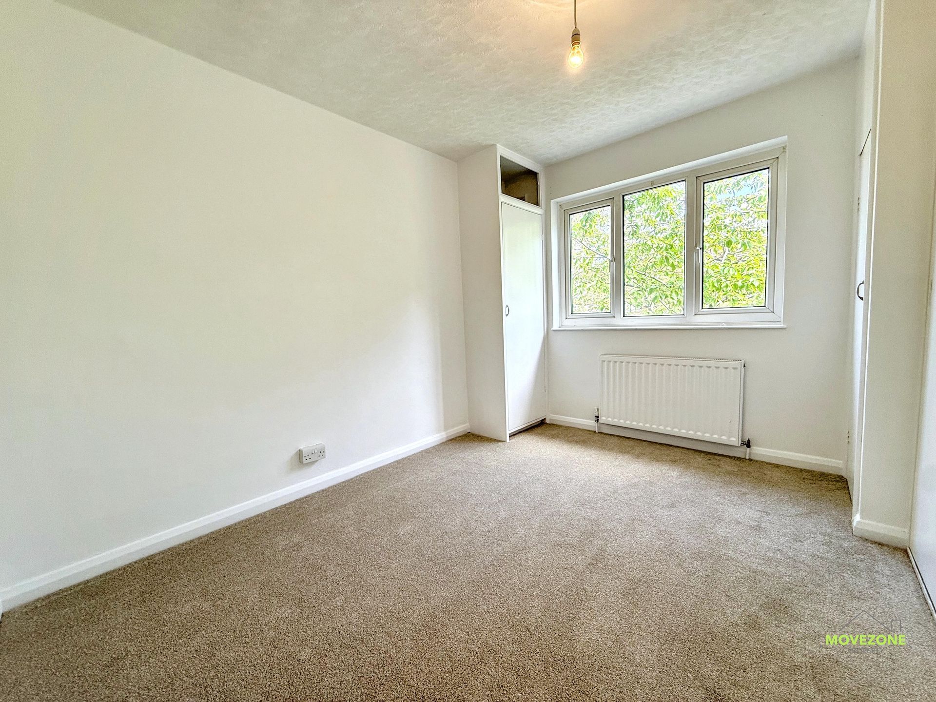 Empty beige-carpeted bedroom with white walls, window, radiator, and a hanging light bulb.