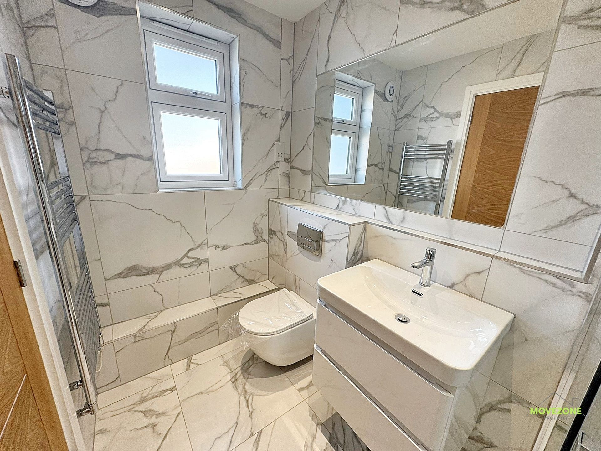 Modern white tiled bathroom with toilet, sink vanity, mirror, shower, and window.