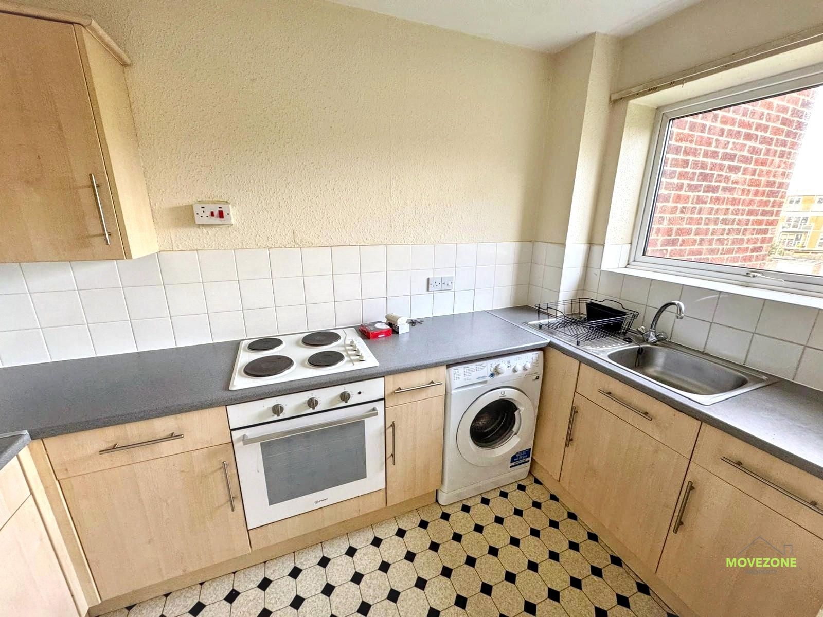Small kitchen with light wood cabinets, gray counters, white stove, washing machine, sink, and checkered floor.