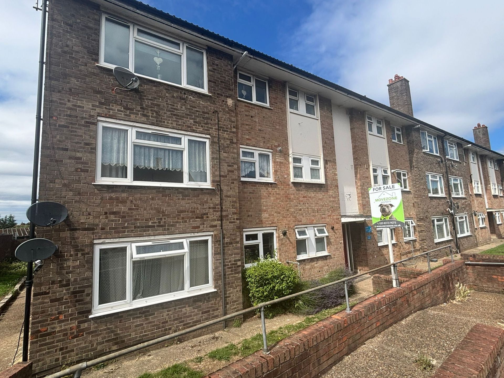 Brick apartment building with white-framed windows and a small front garden under a cloudy sky