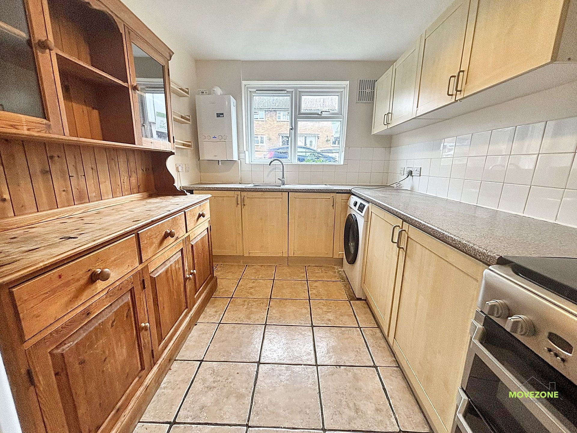 Narrow galley kitchen with wooden cabinets, tiled floor, white appliances, and a window over the sink.