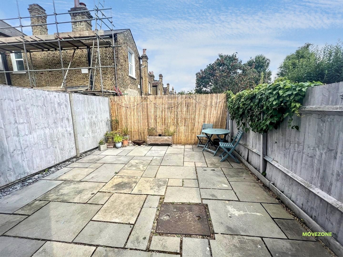 Stone-paved backyard patio with wooden fence, concrete walls, greenery, and a wheelbarrow near the far end