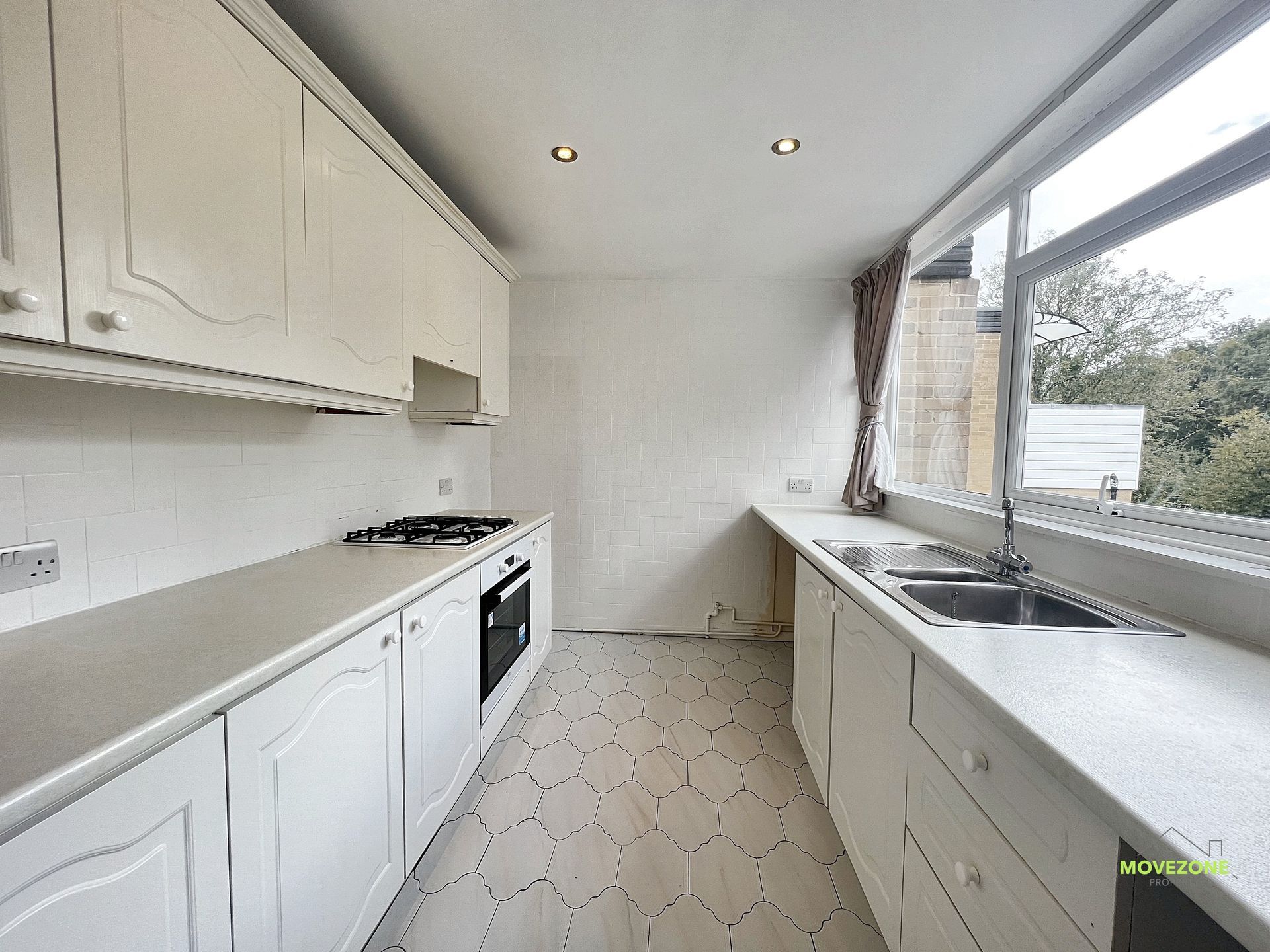 Bright white galley kitchen with cabinets, oven, sink, and large window overlooking greenery