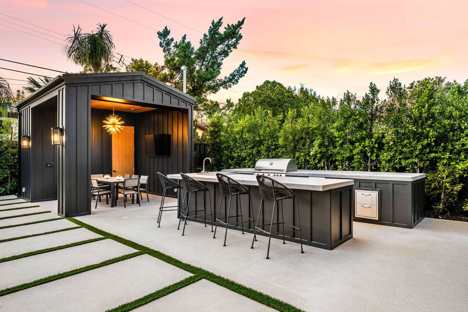 Modern outdoor kitchen and dining patio with dark cabinets, bar stools, and string lights at sunset