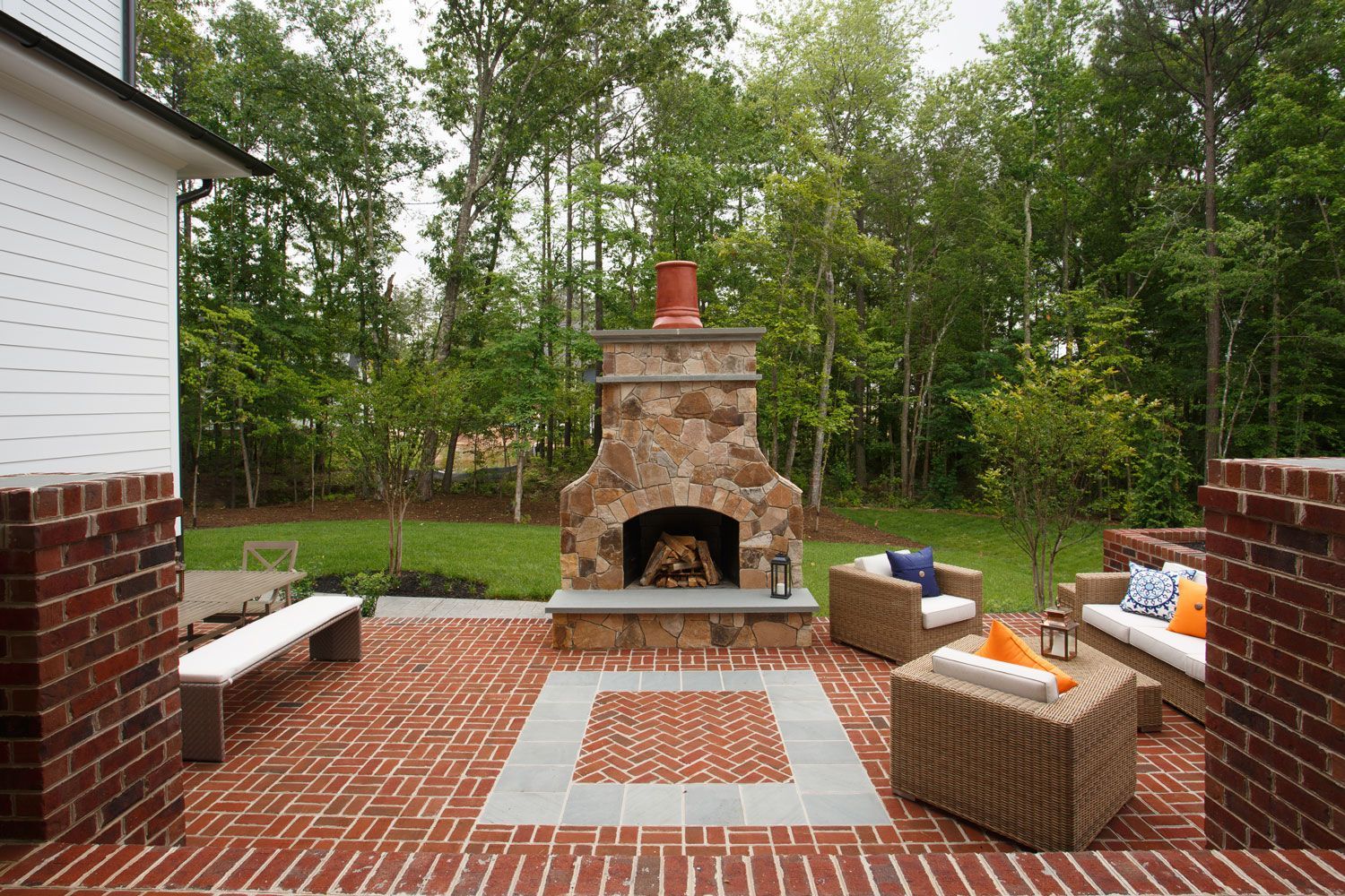 Brick patio with outdoor stone fireplace and wicker seating in a wooded backyard