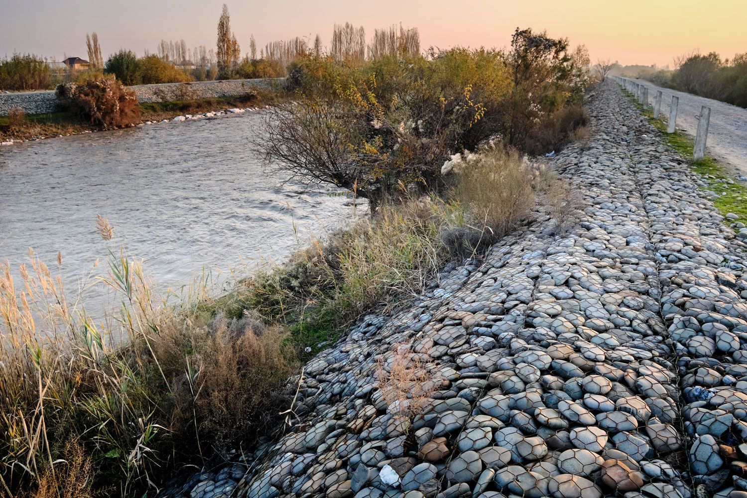 Rocky riverbank beside a calm waterway at sunset, with shrubs and a gravel path along the edge