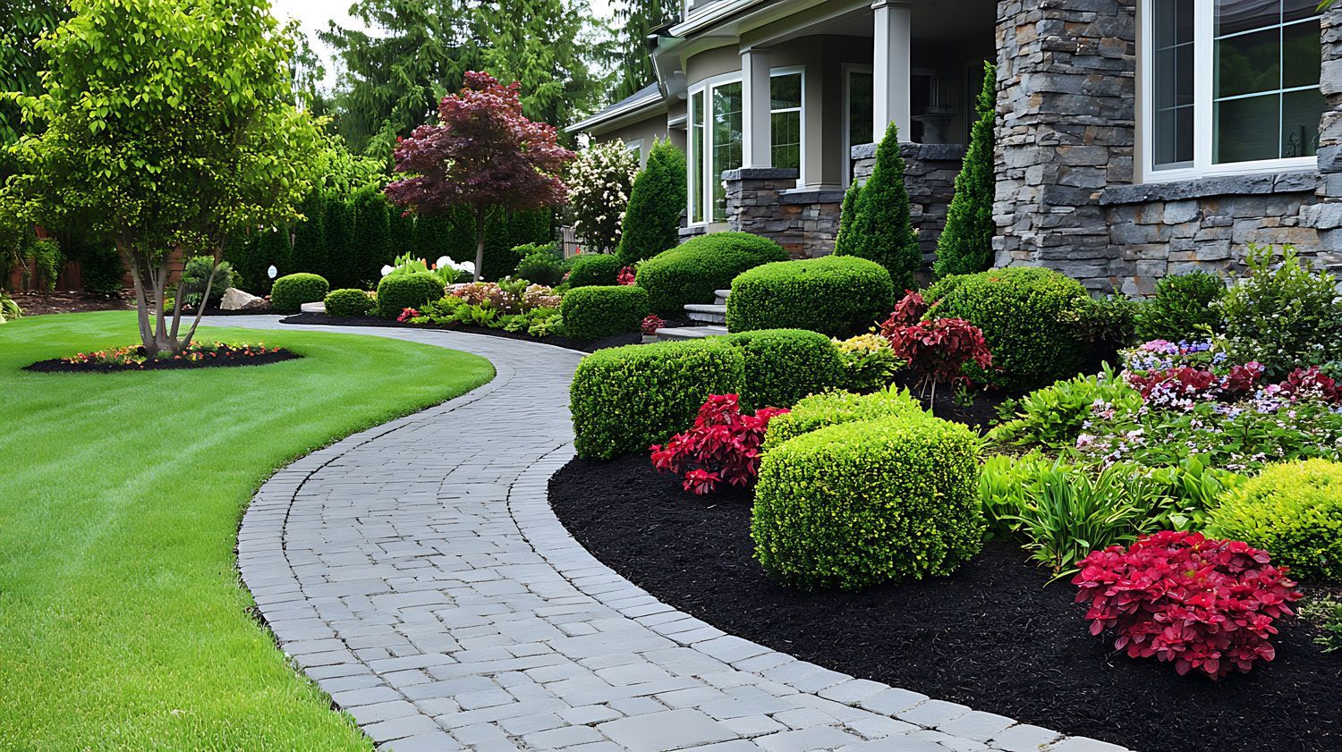 Curved stone walkway beside a house, lined with manicured shrubs, bright flowers, and a green lawn.