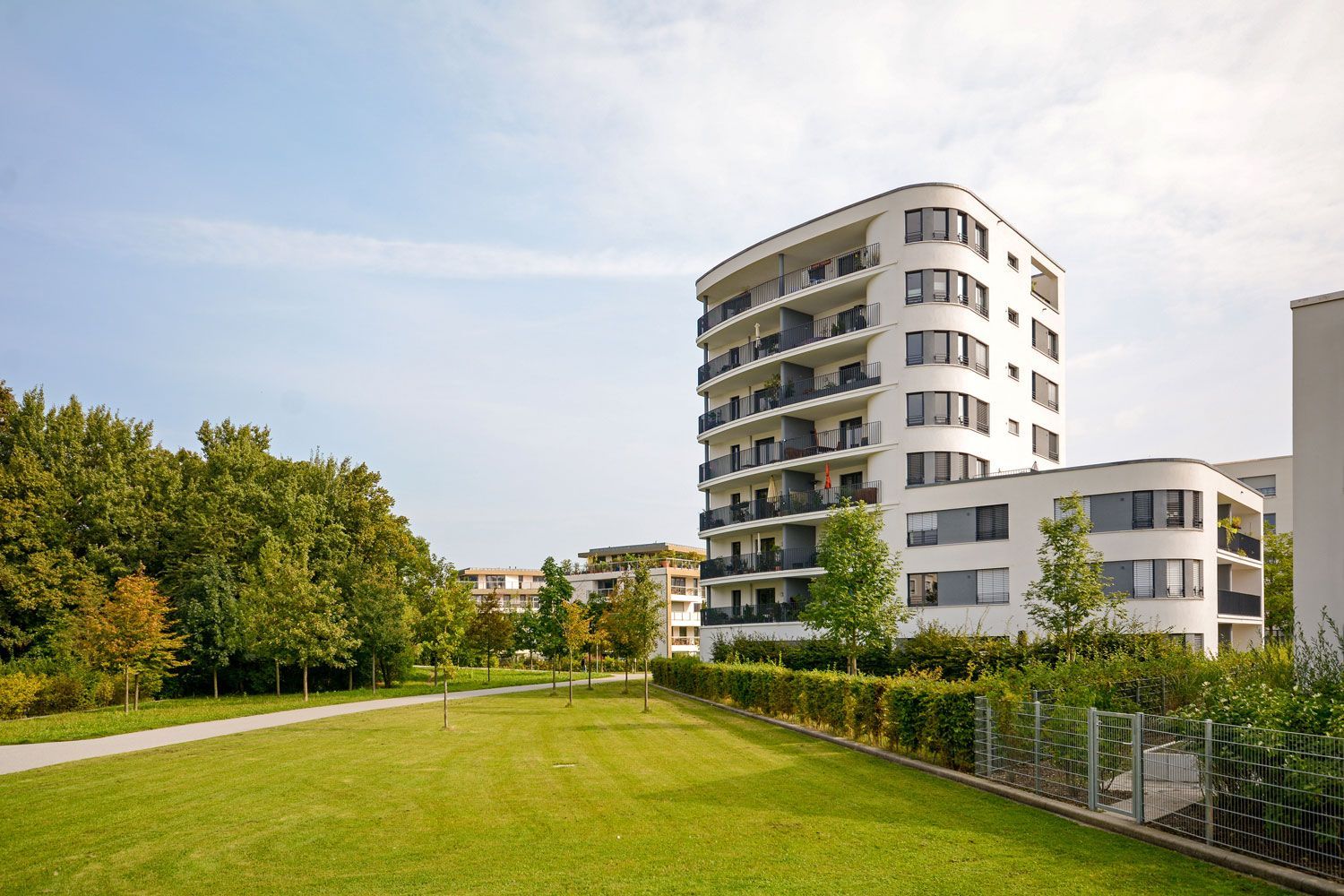 Modern white apartment building beside a green lawn and trees under a partly cloudy sky