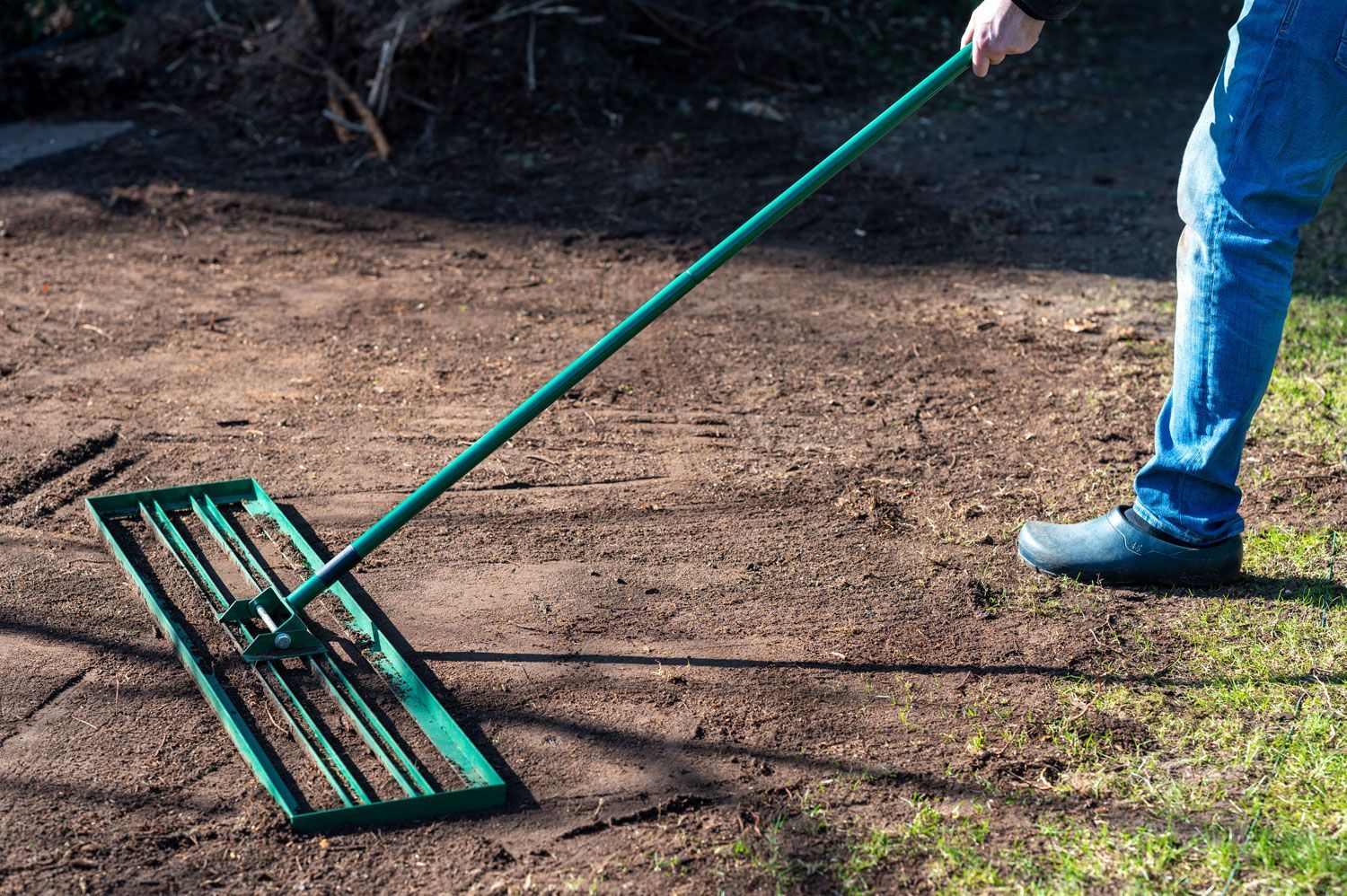 Person raking dirt with a green garden rake on a patchy outdoor yard.