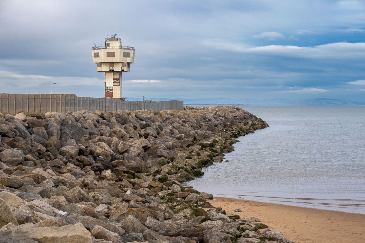 Coastal watchtower on a rocky breakwater beside a calm beach under cloudy sky