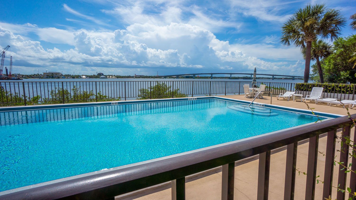 Poolside view of a blue swimming pool overlooking water, with palm trees, lounge chairs, and a cloudy sky