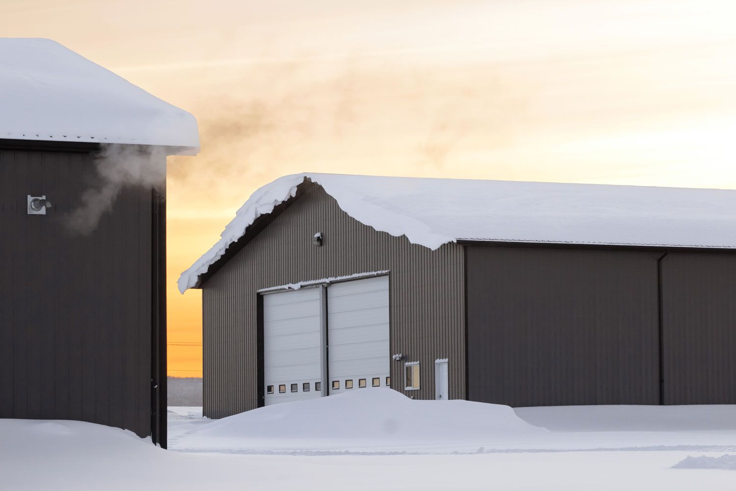 Snow-covered industrial warehouse buildings at sunrise or sunset, with white garage doors and warm sky.