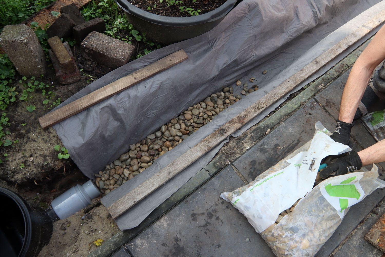 Person installing pebbles in a garden drainage trench with plastic liner and pipes