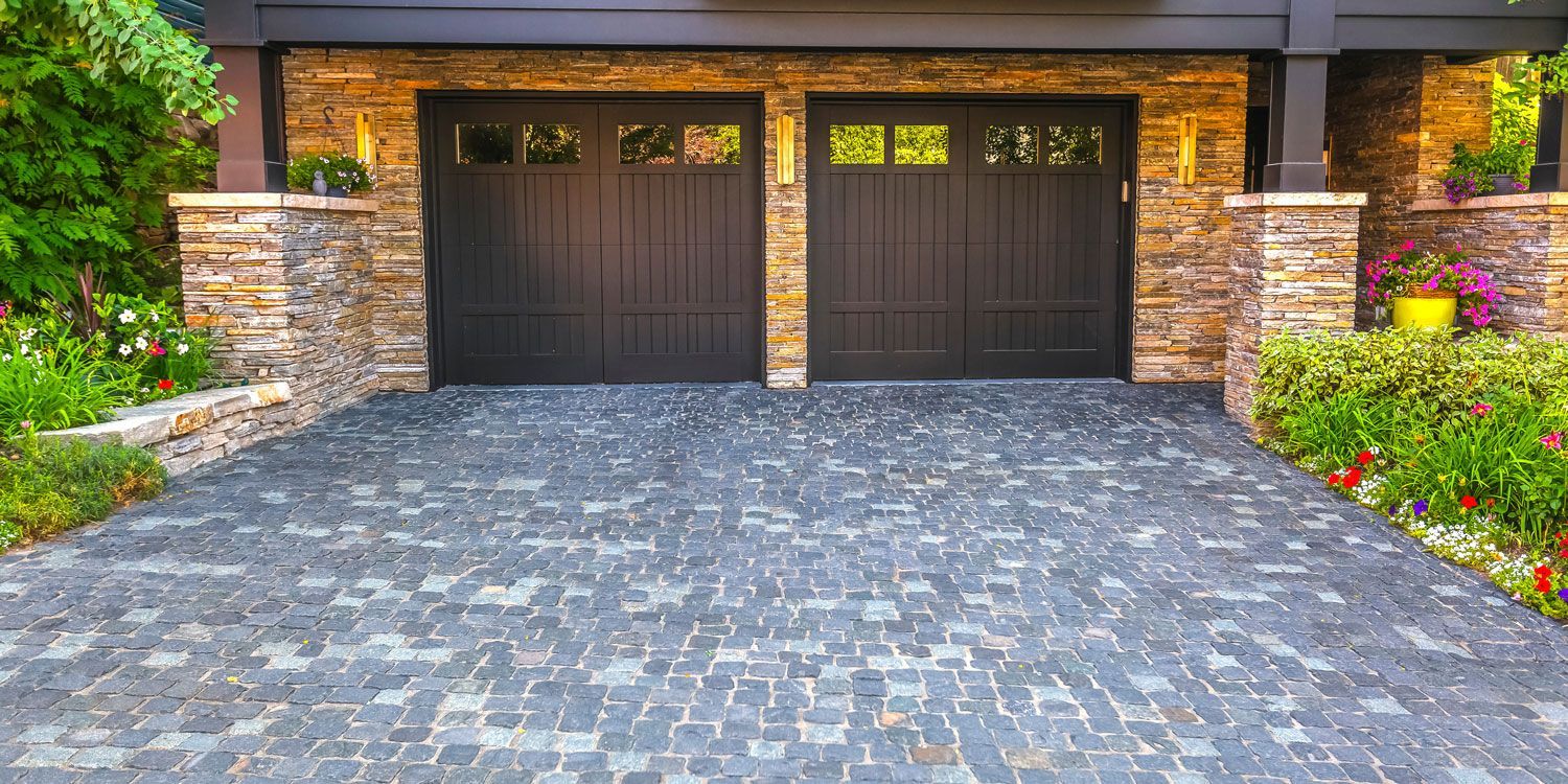 Stone-paver driveway leading to a double garage with dark wooden doors and landscaped flower beds