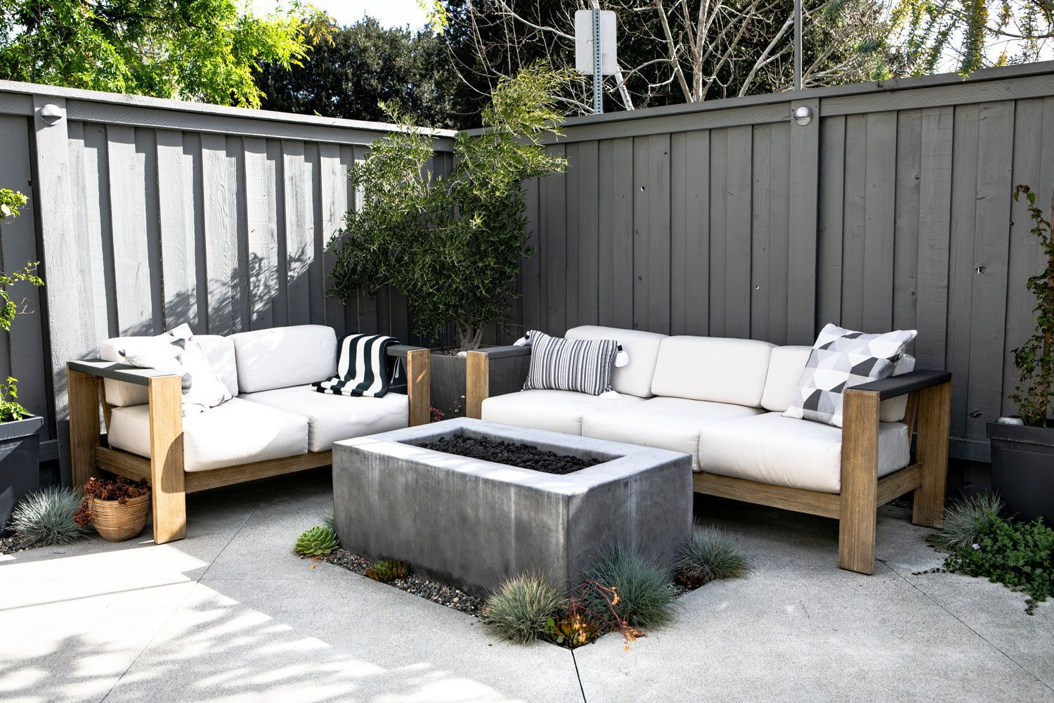 Outdoor patio with two white cushioned benches around a concrete fire pit, against a gray fence.