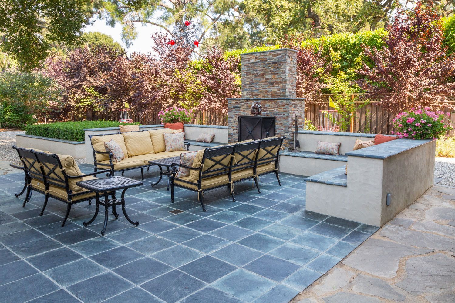 Patio with gray stone pavers, black metal seating, beige cushions, and a stone fireplace surrounded by greenery
