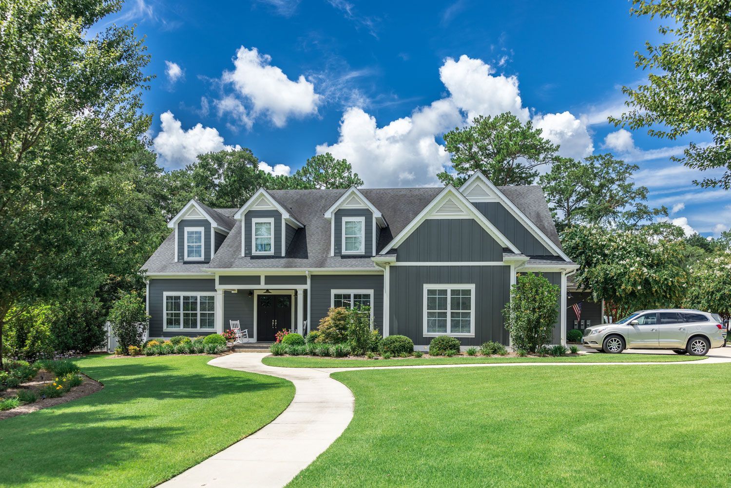 Gray suburban house with white trim, a curved walkway, green lawn, and a parked car under a blue sky