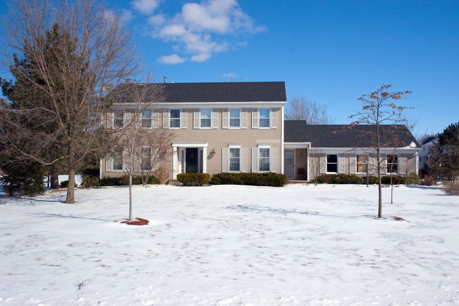 Two-story tan house with dark roof and bare trees in a snowy yard under a blue sky