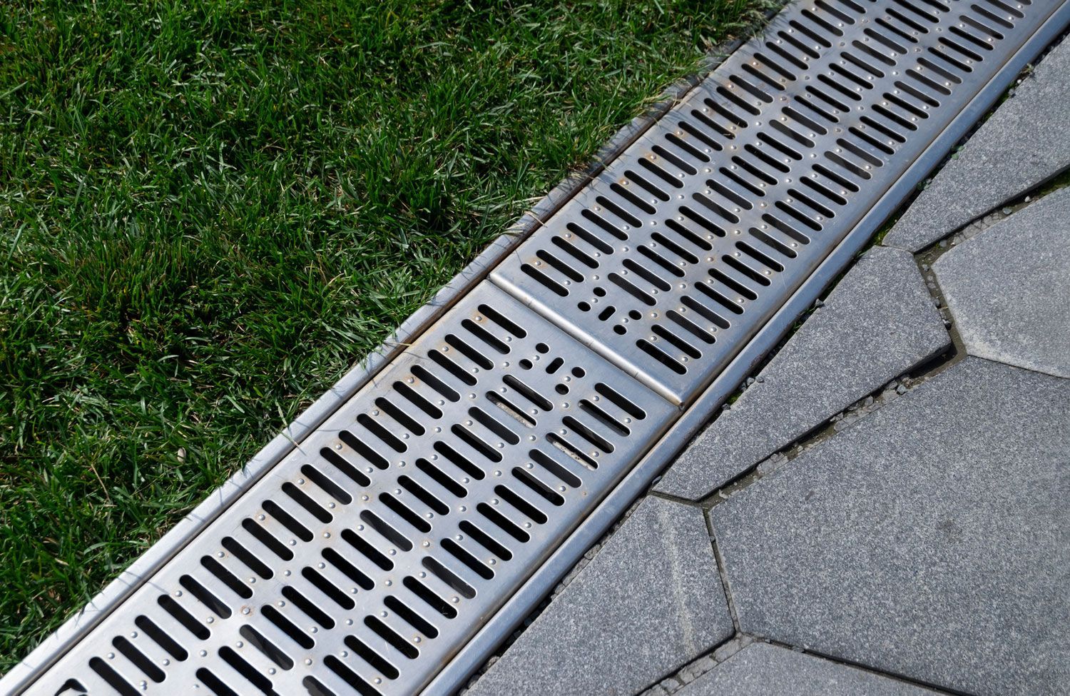 Metal drainage grates between green grass and gray paving stones