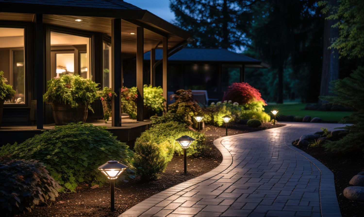 Lit garden path beside a house at dusk, with warm porch lights and landscaped shrubs.
