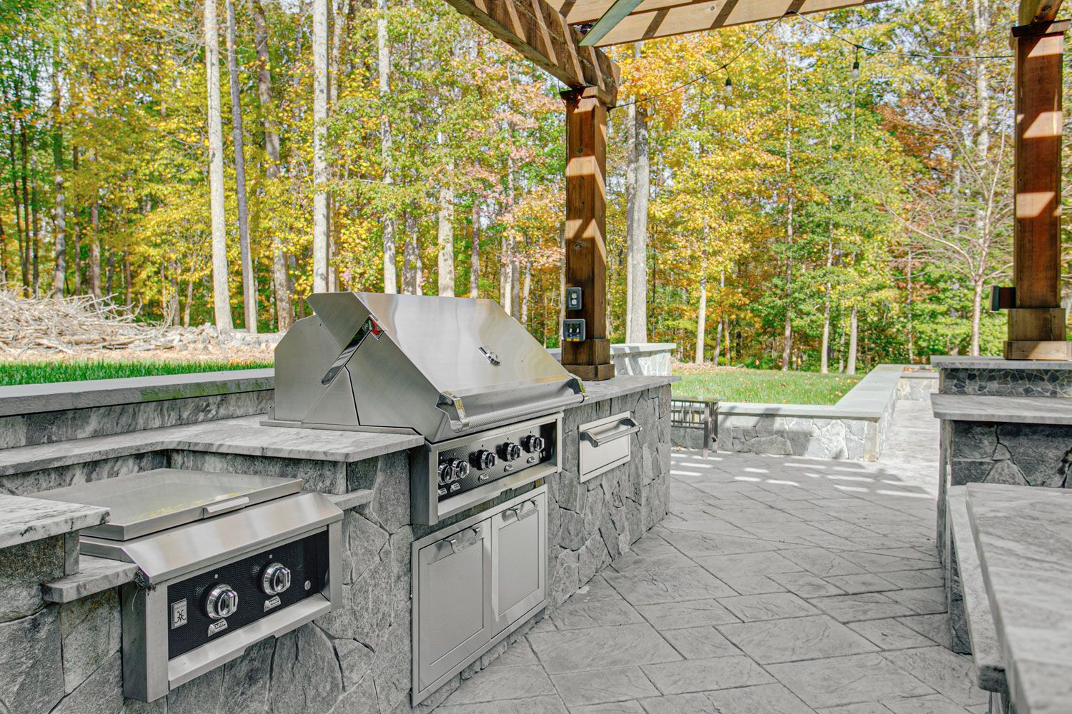 Stone park grill area with built-in barbecue stations under a wooden shelter, surrounded by autumn trees