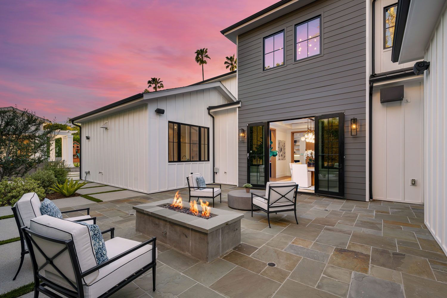 Modern patio with white outdoor seating, fire pit, and large house at sunset