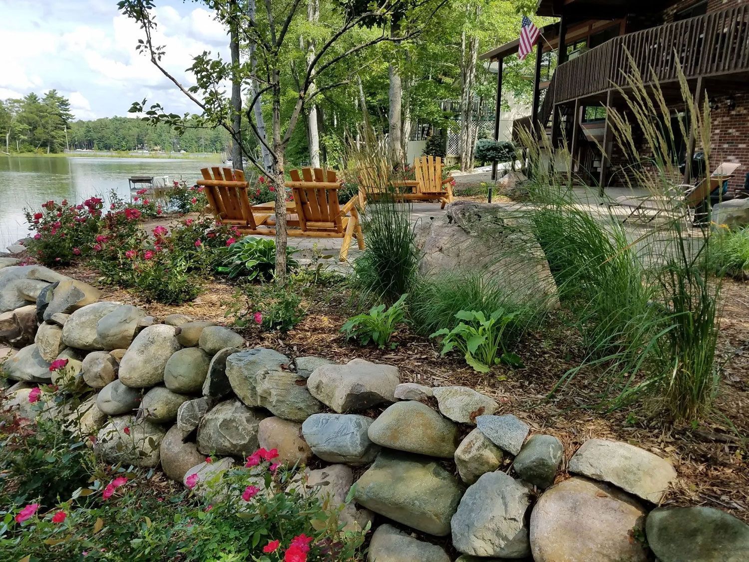 Wooden chairs by a lakeside garden with rocks, flowers, and trees under a rustic cabin deck