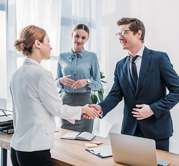 Three businesspeople shaking hands in a bright office, with a laptop and papers on the desk.