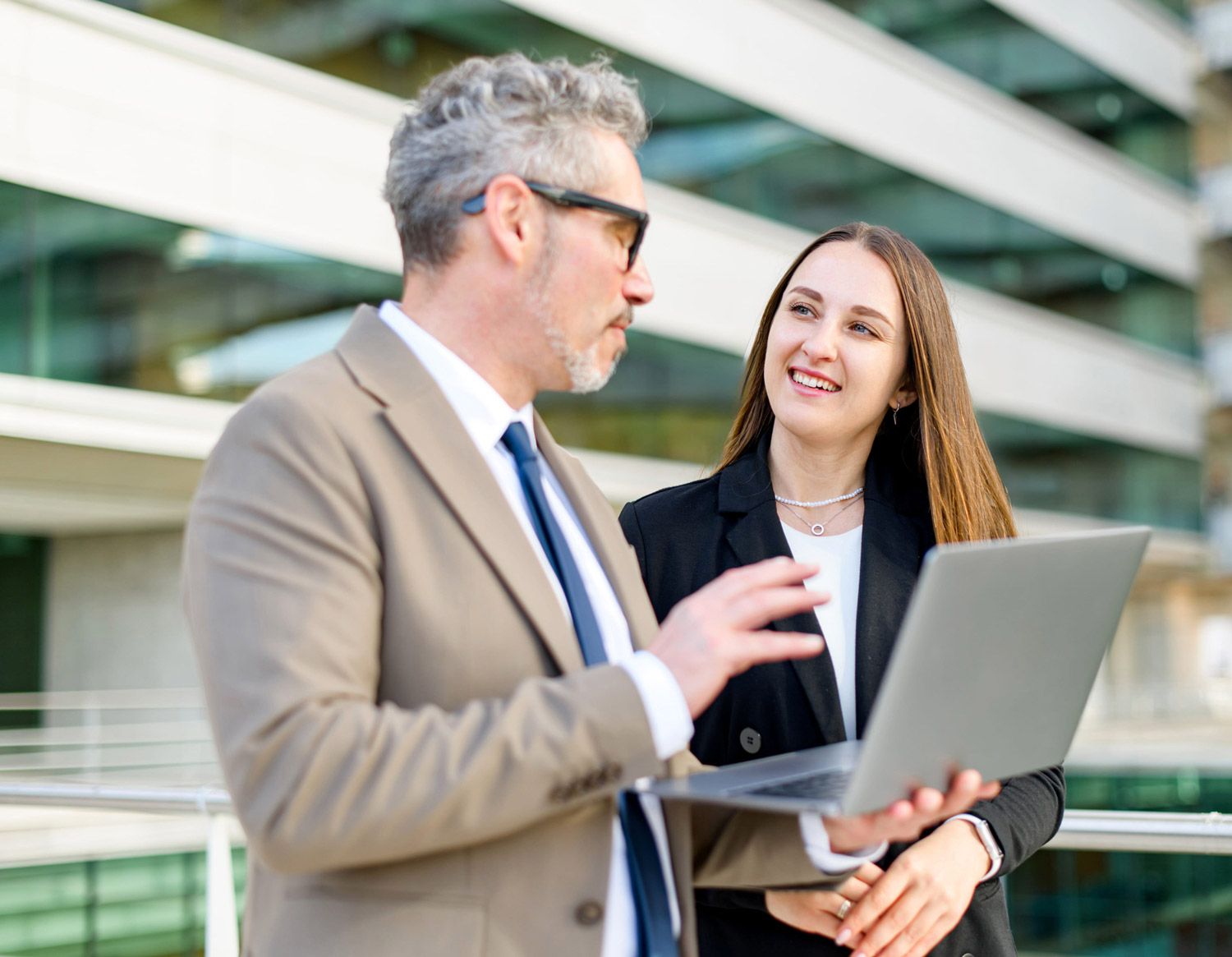 Two businesspeople discussing a laptop outdoors near a modern office building
