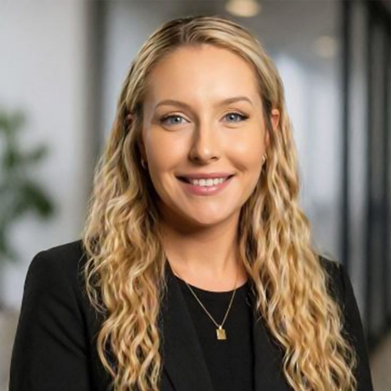 Smiling woman with wavy blonde hair in a black blazer and necklace, standing indoors against a blurred background