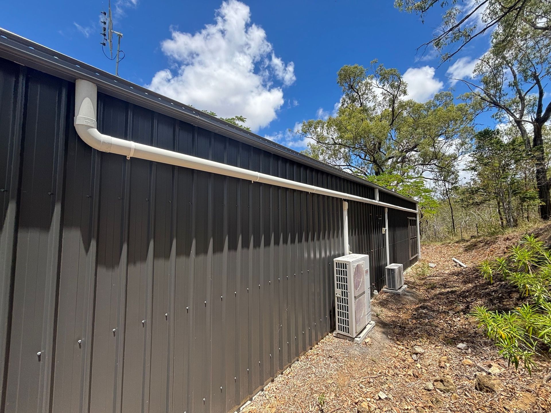 Dark brown building exterior with white gutter and two air-conditioning units along a wooded side yard