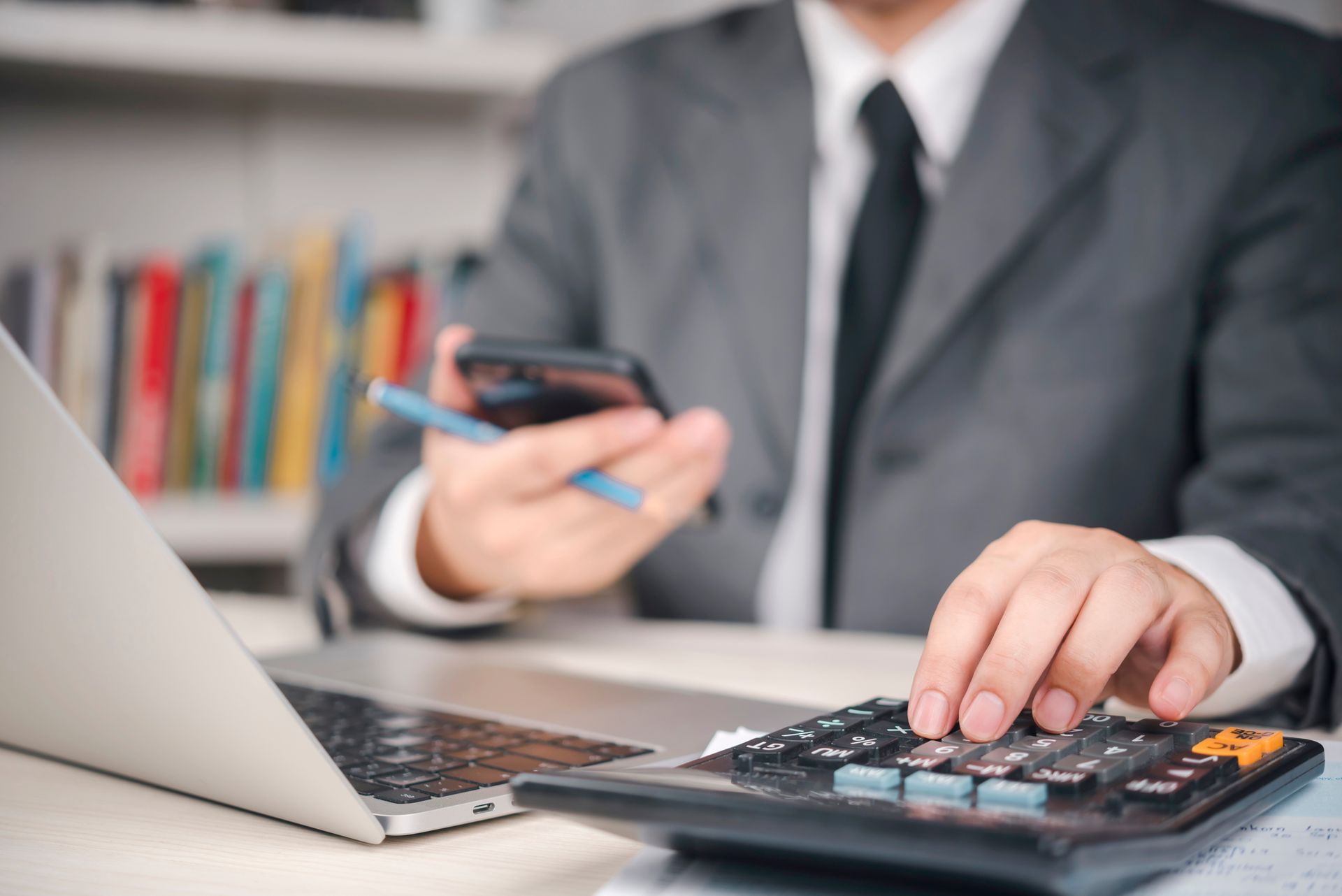 Person in a suit using a calculator, phone, and laptop at a desk.