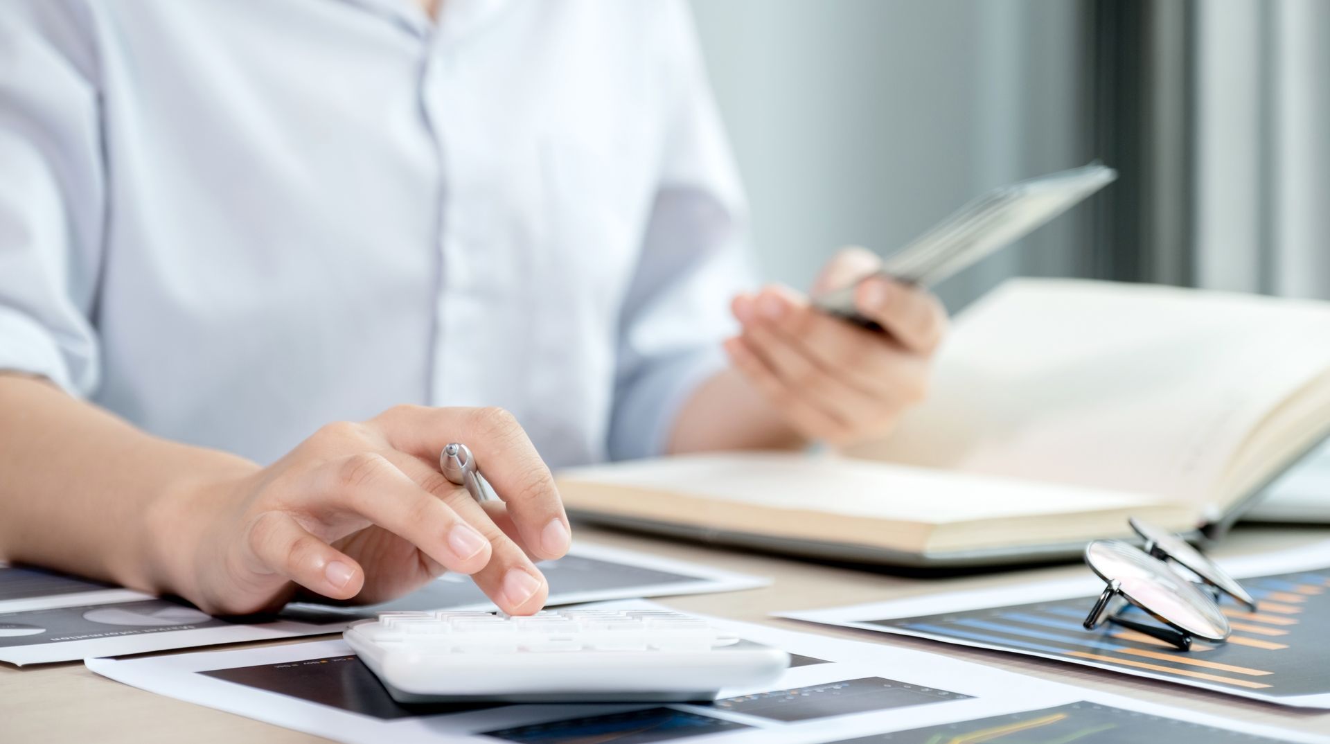 Person using a calculator, holding a tablet and pen, with documents and a notebook on a desk.