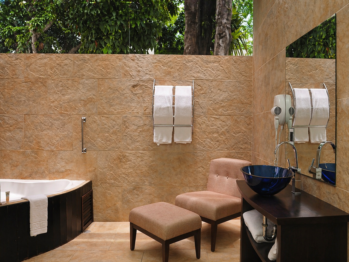 Un baño moderno con bañera, sillón con reposapiés, tocador con lavabo de cristal azul y paredes de azulejos color beige.