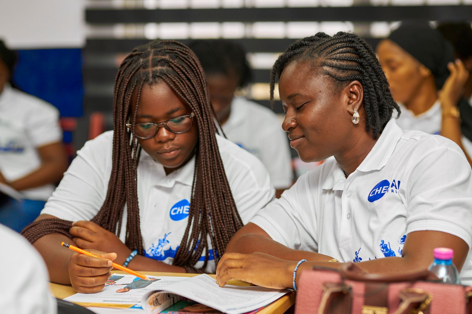 Deux femmes étudient ensemble, l'une d'elles désignant un cahier. Toutes deux portent des chemises blanches à logo et semblent concentrées.