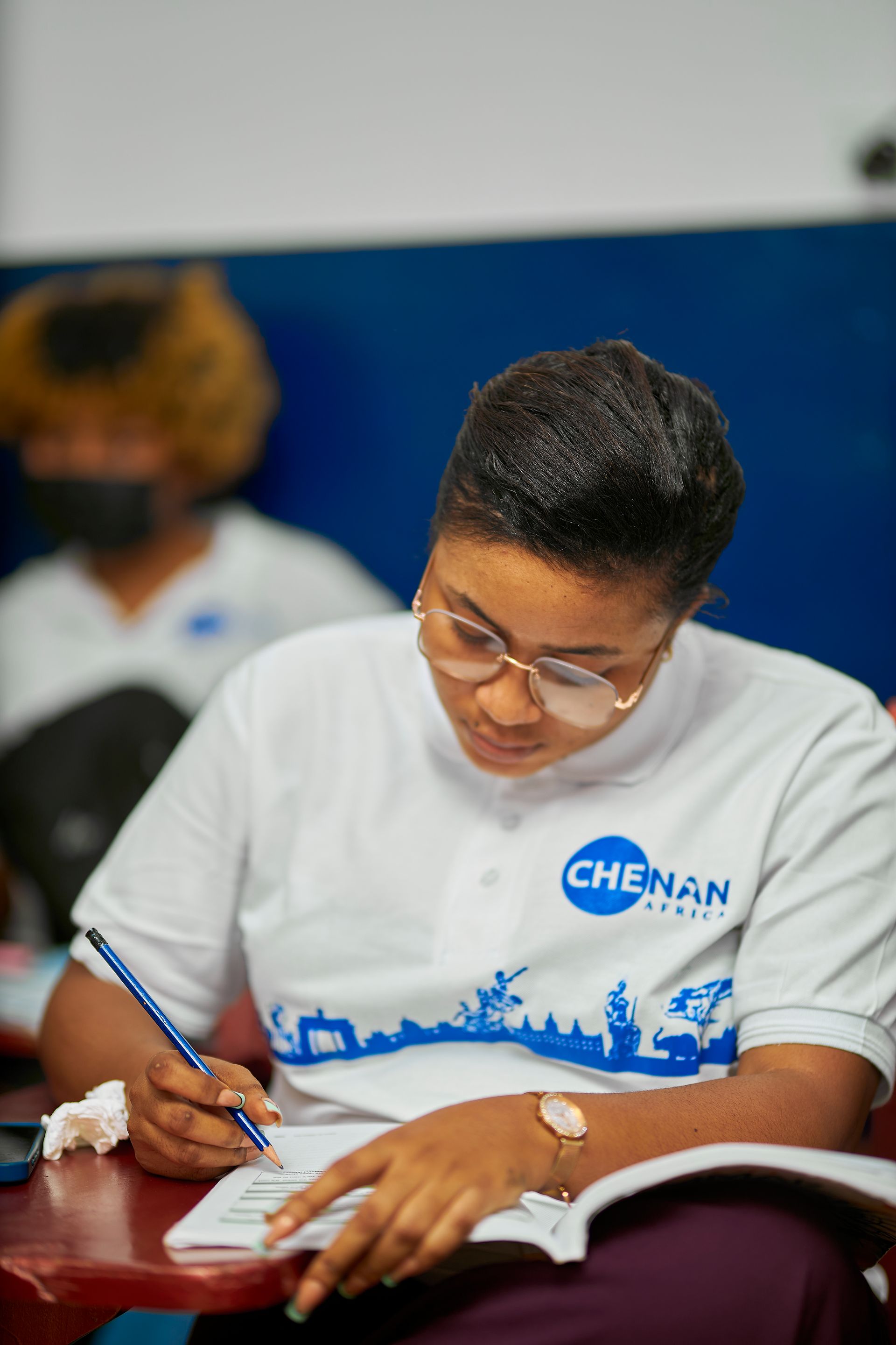 Femme à lunettes écrivant dans un carnet, vêtue d'une chemise blanche avec le logo « CHENAN ».