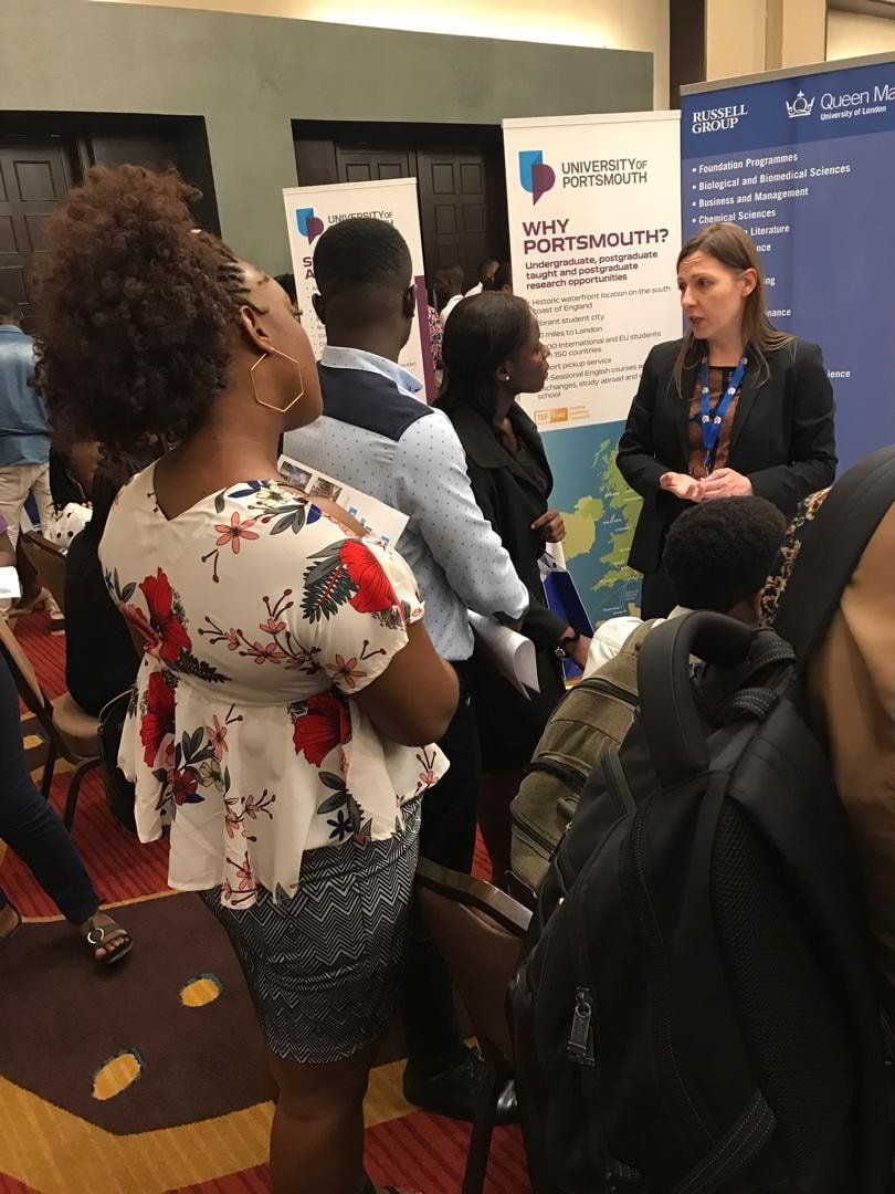 Des personnes assistent à une conférence. Une femme prend la parole sur un stand entouré de plusieurs participants. Des affiches et des cartes sont visibles en arrière-plan.