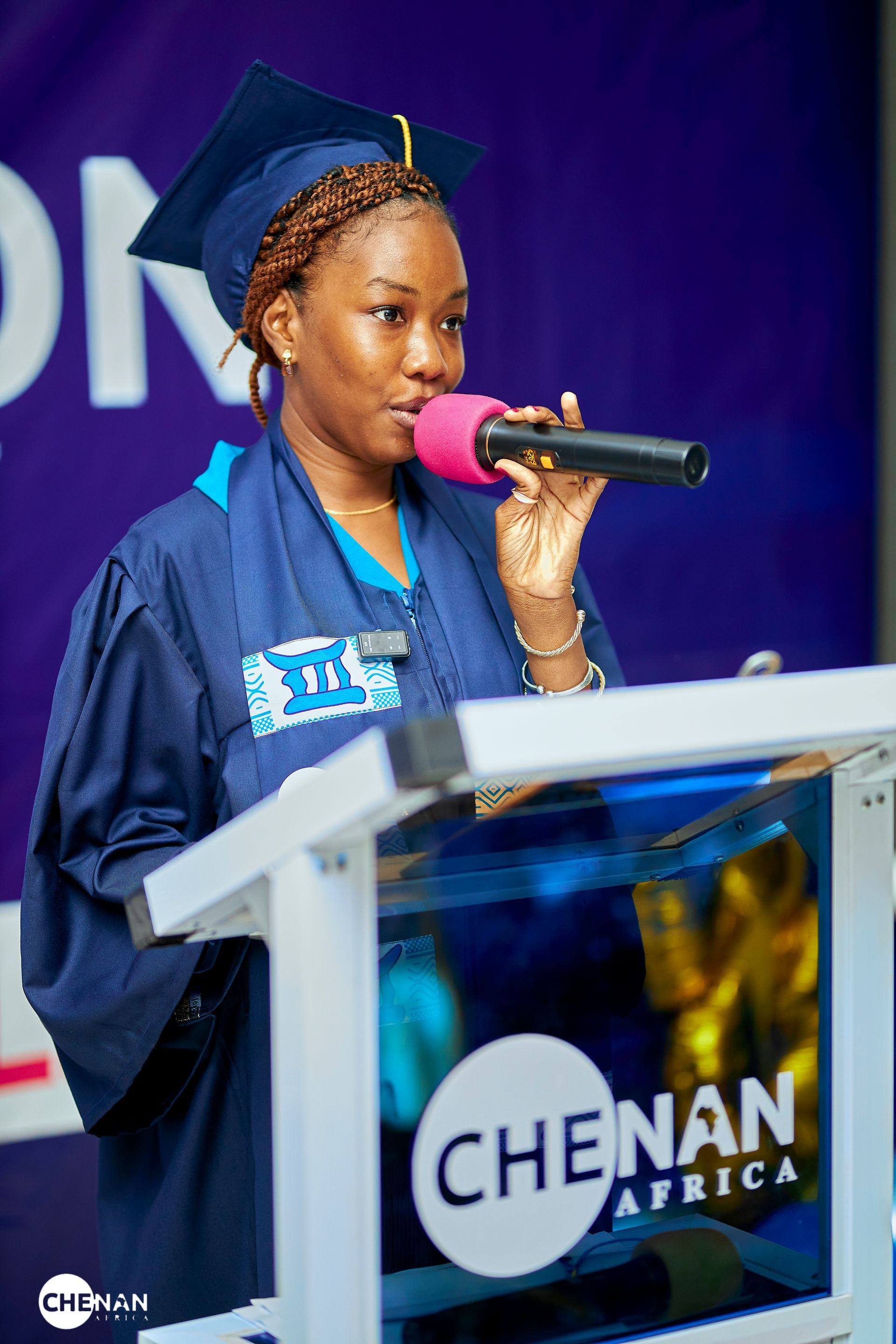 Une femme en robe de remise de diplôme prend la parole au micro, sur un podium orné du logo « CHENAN AFRICA ».